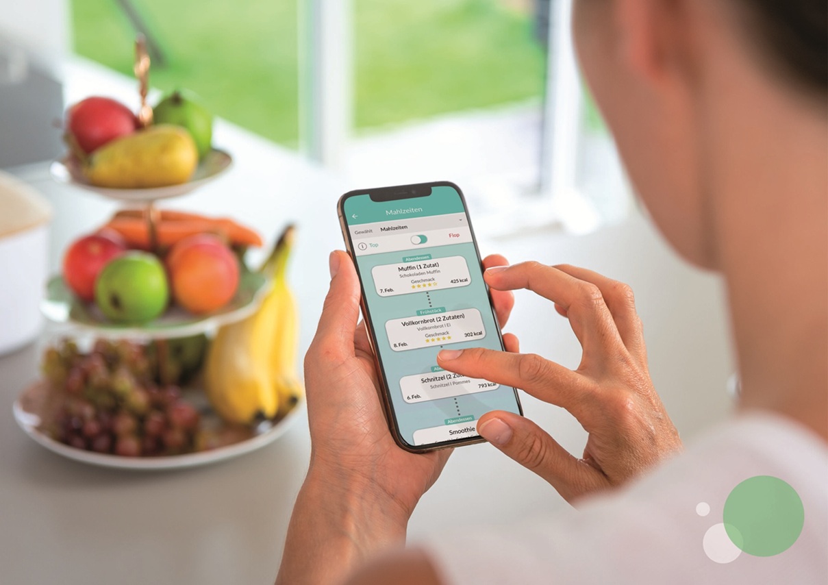Eine Person bedient ihr Handy, Blick über die Schulter. Im Hintergrund ist Unscharf ein Etagere mit Obst zu erkennen.